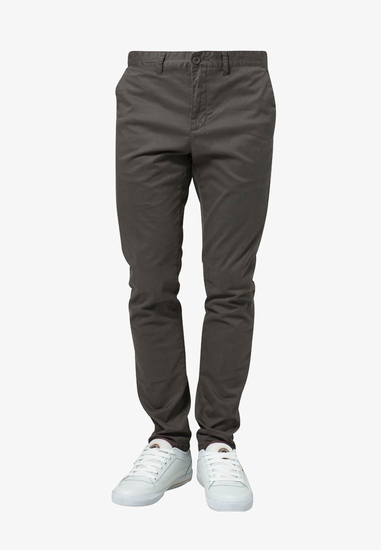 Pantalon gris foncé ajusté associé à des baskets blanches basses portées par une personne se tenant devant un fond blanc.