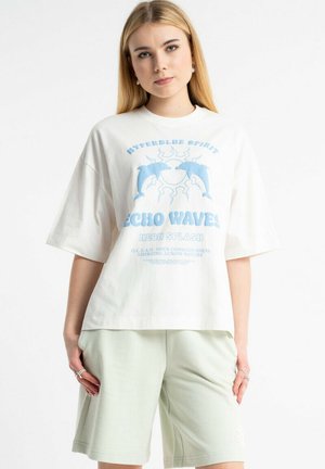 Młoda kobieta w białym, oversize T-shircie z niebieskim delfinem i słońcem oraz jasnooliwkowymi szortami stoi na białym tle.