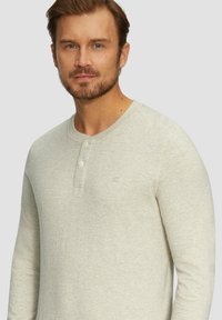 Helles graues, langärmliges Henley-Shirt aus weichem Material, mit einem runden Kragen und drei Vorderknöpfen. Dezentes Logo-Detail sichtbar.