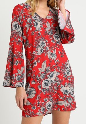 Femme portant une robe rouge à fleurs avec des manches longues évasées et un décolleté en V. Le motif floral est noir et blanc.
