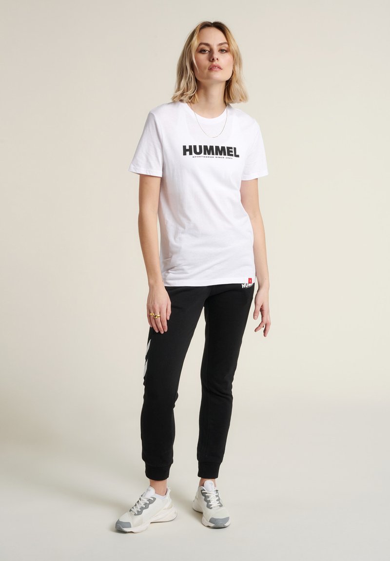 T-shirt en coton blanc avec "HUMMEL" imprimé en noir. Associé à un pantalon de jogging noir avec des accents d'éclairs blancs et des baskets grises.