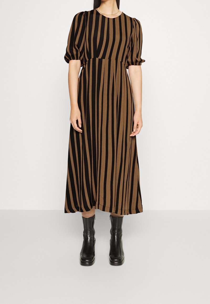 Femme portant une robe midi à rayures verticales marron et noire avec des manches bouffantes et des bottines noires, debout sur un fond blanc.