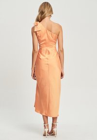 Robe en satin à une épaule en pêche avec un accent en nœud à l'épaule. Longueur midi et texture lisse, avec un corsage ajusté et de subtils plis.