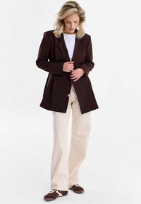 Hnedé sako s výrezovým klopou, v kombinácii s bielou košeľou a krémovými nohavicami, vybavené voľným strihom a jemným ružovým štítkom.