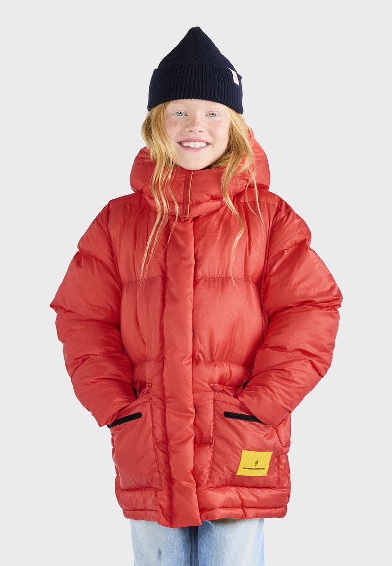 Rote Daunenjacke mit Kapuze, mit einem Reißverschluss vorne, seitlichen Taschen mit schwarzem Besatz und einem gelben Markenlabel. Getragen mit einer marineblauen Strickmütze.