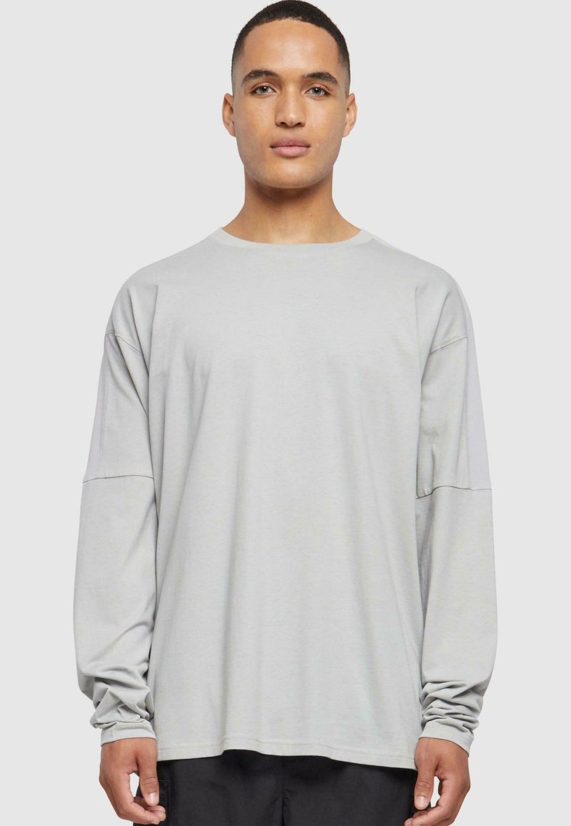 Lys grå langermet skjorte laget av mykt stoff, med bred halsutskjæring, senkede skuldre og en avslappet passform. Glatt tekstur, minimale sømmer.