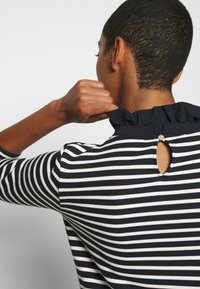 Raidallinen paita, jossa on tummansinisiä ja valkoisia vaakaraidoituksia, röyhelökaulus, avainreikäyksityiskohta takana ja kolme neljännestä hihoista.