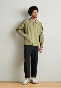 Olive green button-up jacket with a single pocket and logo, paired with black textured pants and green and cream sneakers.
