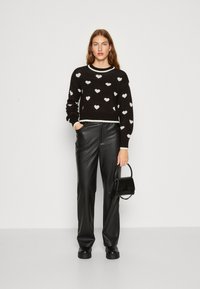 Črn skrajšan pulover z belimi srčkastimi vzorci, združen z črnimi usnjenimi hlačami in majhno črno torbo, ki ima strukturirano zasnovo.