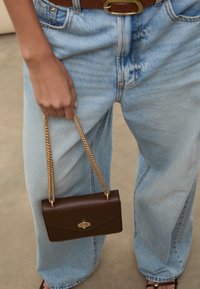 Smeđa kožna torba s naramenicom od lanca, s prednjim preklopom i zlatnim ukrasima, drži se uz svijetloplave široke traperice.