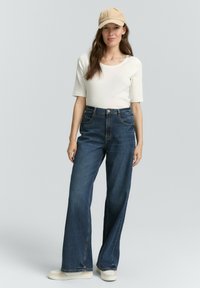 Lichtbeige pet, witte T-shirt met korte mouwen, wijde donkerblauwe jeans en crèmekleurige schoenen. De stof heeft een gladde textuur met minimale details.