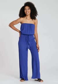 Mono azul sin mangas hecho de una tela ligera; cuenta con un corsé fruncido, cintura con cordón ajustable y piernas anchas y fluidas con acentos de borlas.