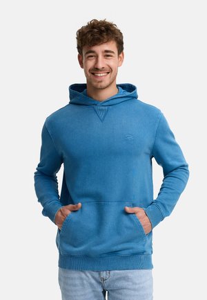 Smilende mand med kort krøllet hår iført en blå hættetrøje med hænderne i lommen, stående mod en ensfarvet baggrund.