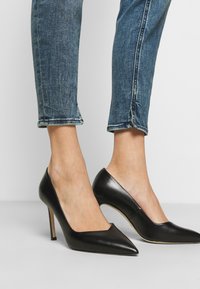 Zapatos de tacón alto negros se combinan con jeans cortos azules, destacando la punta afilada y el diseño elegante sobre un fondo neutro.