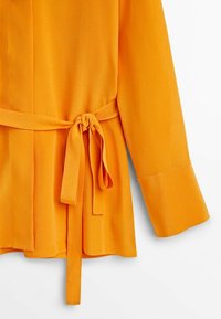 Blouse orange à manches longues avec larges poignets et ceinture en tissu nouée en nœud à la taille sur fond blanc.