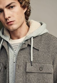 Harmaa tekstiilitakki, jossa on paitakaulus, mukautuva beige fleece-vuorattu huppu, etupainikkeet ja harmaa vetoketjuhuppari alla.