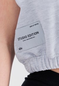Svetlo šedý top s krátkymi rukávmi a nariaseným spodkom, so štítkom s nápismi "STUDIO EDITION" a "ALPHA INDUSTRIES INC." čiernym písmom.