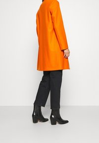 Orange uldfrakke med lige snit, lange ærmer og en ren silhuet. Kombineret med sorte denimbukser og blanke ankelstøvler med hæl.