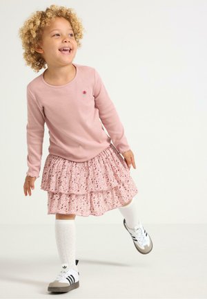 Ljusrosa långärmad topp med rosbroderi, kombinerad med en rosa blommig volangkjol. Vita knästrumpor och svarta sneakers med vita accenter.