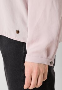 Vaaleanpunainen paita, jossa on sileä pinta, pyöreä helma ja tummat napit hihoissa, yhdistettynä tummanharmaisiin housuihin.