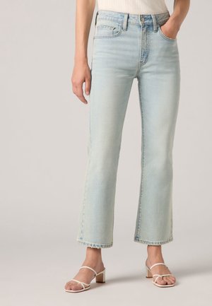 Vrouw die lichtblauwe cropped flare jeans en witte sandalen met blokhak en bandjes draagt, met één hand in de zak, tegen een effen achtergrond.