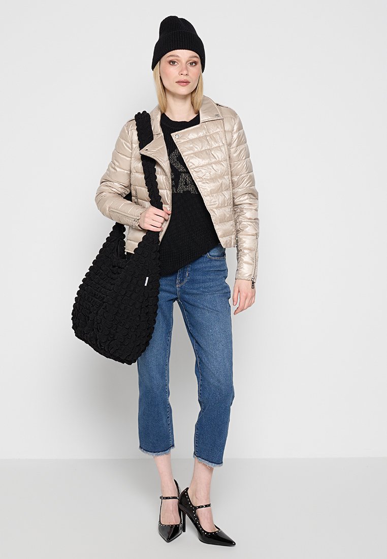 Beige Steppjacke, schwarzer Strickpullover, verkürzte blaue Jeans, schwarze strukturierte Tragetasche und schwarze lackierte High Heels mit Nietenakzenten.