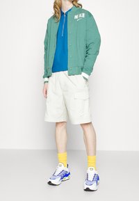 Grön bomberjacka med vita detaljer, blå pikétröja, beige cargoshorts, gula strumpor och flerfärgade sneakers.