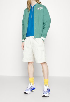 Veste bomber verte avec des accents blancs, chemise polo bleue, shorts cargo beiges, chaussettes jaunes et baskets multicolores.