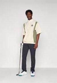 Top de golfe bege e verde-oliva de mangas curtas com gola alta, combinado com calças escuras e sapatos de golfe brancos. Segura um taco de golfe.