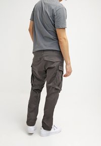 Pantalon cargo gris avec plusieurs poches latérales, se resserrant vers les chevilles, assorti à un t-shirt gris à manches courtes et des baskets blanches.