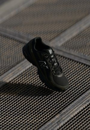 Schwarzer New Balance Sneaker mit Schnürsenkeln auf Metallgitteroberfläche, schräg, um Seiten- und Frontdetails zu zeigen.