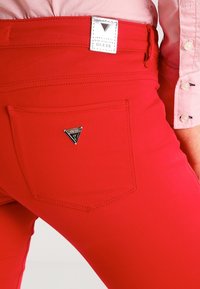 Røde cowboybukser i denim med en baglomme, der har et trekantet Guess-logo i sølv, fremhævet af rene sømme og en strømlinet pasform.