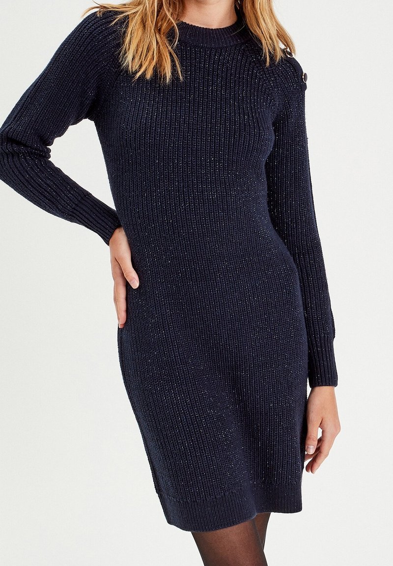 Femme portant une robe pull ajustée côtelée bleu marine à manches longues avec des détails de boutons sur l'épaule, debout avec une main sur la hanche.