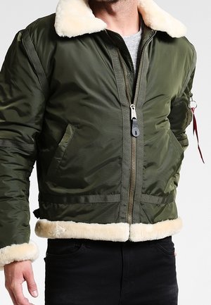 Mężczyzna noszący zieloną kurtkę typu bomber z beżowym kołnierzem, mankietami i obrębem z baranka, częściowo rozpiętą na szarej koszuli.