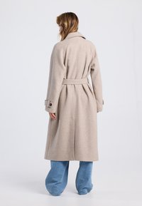 Abrigo de mezcla de lana beige con cintura ceñida por un cinturón, cuello ancho y mangas largas, combinado con pantalones de pierna ancha de tono azul claro.