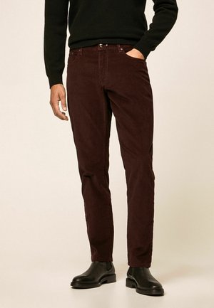 Uomo con pantaloni di velluto a coste rosso scuro, maglione nero, stivali Chelsea in pelle nera, in piedi con una mano in tasca.