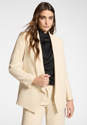 Donna con lunghi capelli castani, indossa un completo color crema e una camicetta nera con bottoni, in piedi davanti a uno sfondo chiaro e uniforme.