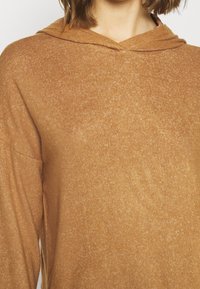 Felpa con cappuccio marrone realizzata in un tessuto texture, leggermente slubbato. Presenta un profondo scollo e maniche dalla vestibilità rilassata. Nessun motivo o accento visibile.