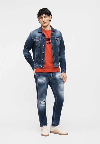 Jeune homme portant une veste en jean déchirée bleu foncé et un jean, un t-shirt rouge à motif, et des baskets blanches, debout devant un fond uni.