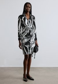 Sort-hvitt marmorert kjole med lange ermer og en tettsittende silhuett, kombinert med en liten svart veske og spisse flate sko.