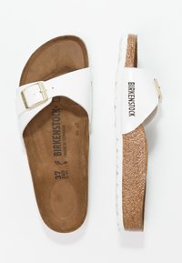 Witte leren schuifsandalen met een verstelbare gouden gesp, kurken voetbed en een gestructureerde rubberen zool. Merkwaarde "BIRKENSTOCK" gedrukt op de zijkant.