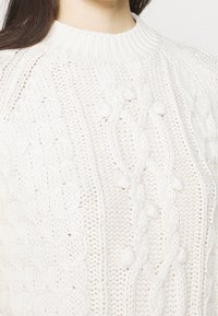 Vit stickad tröja med texturerade kabelmönster och upphöjda bubbel detaljer. Klassisk rund halsringning, medelviktsmaterial och mjuk struktur.