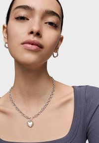 Collar de cadena de plata con un colgante en forma de corazón, usado sobre un top morado texturizado. La modelo lleva aretes de aro y tiene la piel suave.