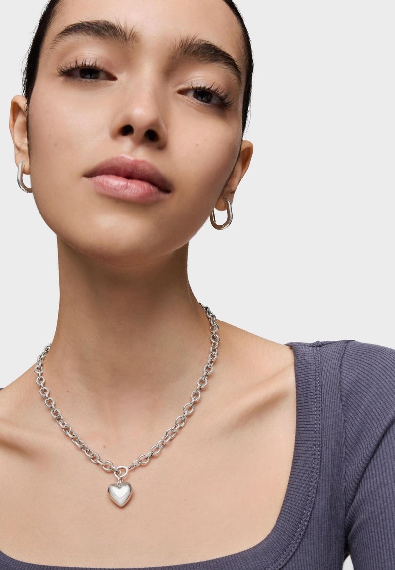 Collar de cadena de plata con un colgante en forma de corazón, usado sobre un top morado texturizado. La modelo lleva aretes de aro y tiene la piel suave.