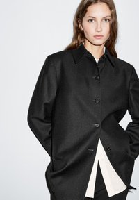 Cappotto in misto lana nera con colletto classico, chiusura a bottoni e due tasche laterali. Abbinato a una camicia bianca con finitura texture.