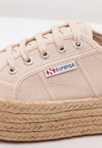 Sneaker in canvas beige con suola piatta in juta intrecciata. Presenta occhielli in argento e un'etichetta Superga rossa/blu sul lato.