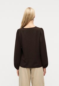 Γυναίκα με μακριά ξανθά μαλλιά φοράει μπλούζα μακριάς μανίκας σκούρου καφέ χρώματος με φουσκωτούς ώμους και ανοιχτό μπεζ παντελόνι με φαρδιά πόδια, στέκεται γυρισμένη προς τα πίσω.