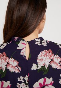 Květinová halenka s tmavě modrým pozadím, zdobená růžovými a bílými květy. Má klíčový výřez na zadní straně, který je upevněn černým knoflíkem.