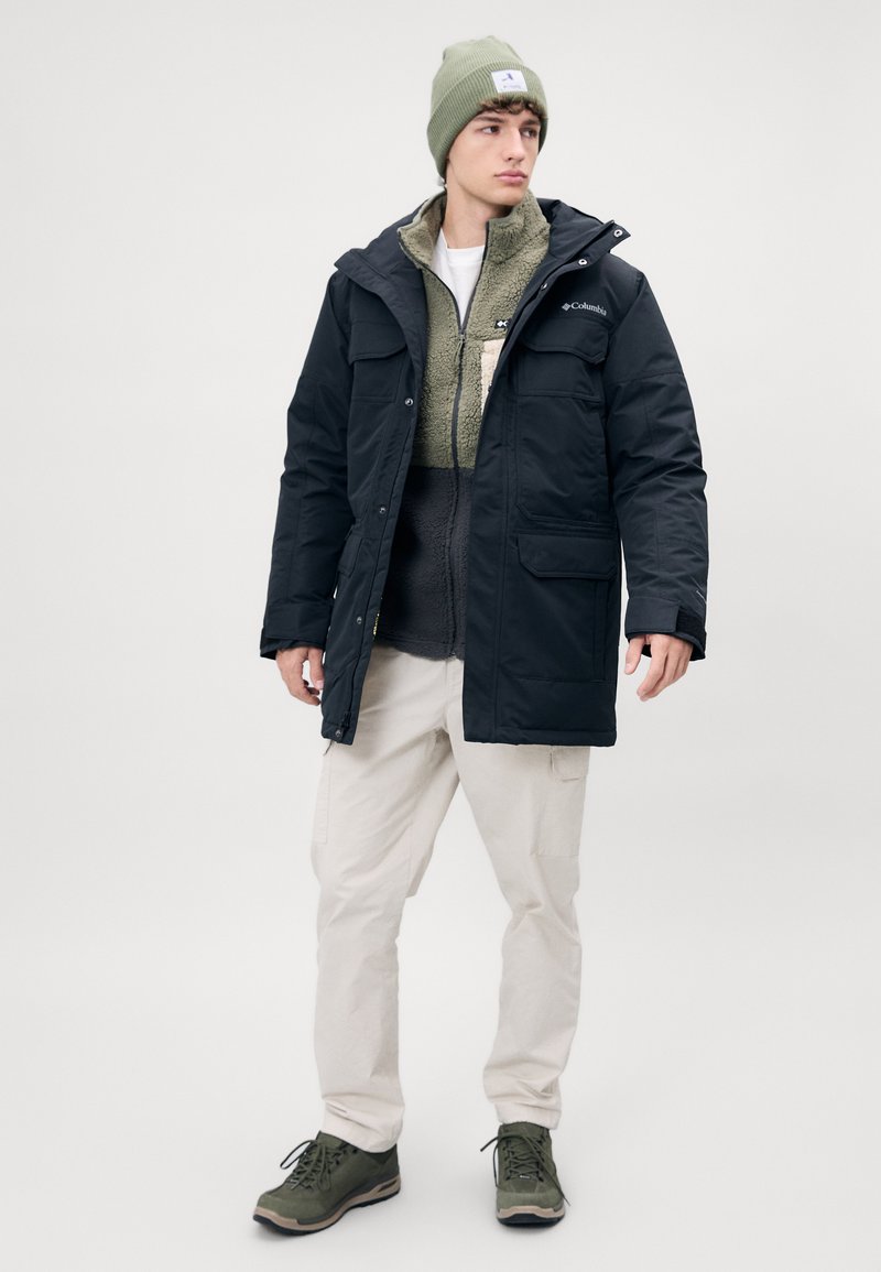 Zwarte waterdichte jas met capuchon, tan fleece voering, beige cargobroek, groene wandelschoenen en een lichtgroene gebreide muts.