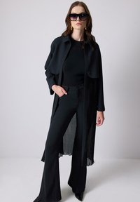 Black, layered outfit featuring a flowing long jacket, fitted top, and flared pants. Glossy heels and oversized sunglasses complete the look.